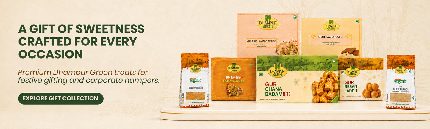 A promotional banner for Dhampur Green treats, showcasing multiple packaged snack boxes and bags arranged on a white pedestal against a light beige background. The packaging features warm tones of orange, yellow, and green, with text overlays on the left reading 'A GIFT OF SWEETNESS CRAFTED FOR EVERY OCCASION'.
