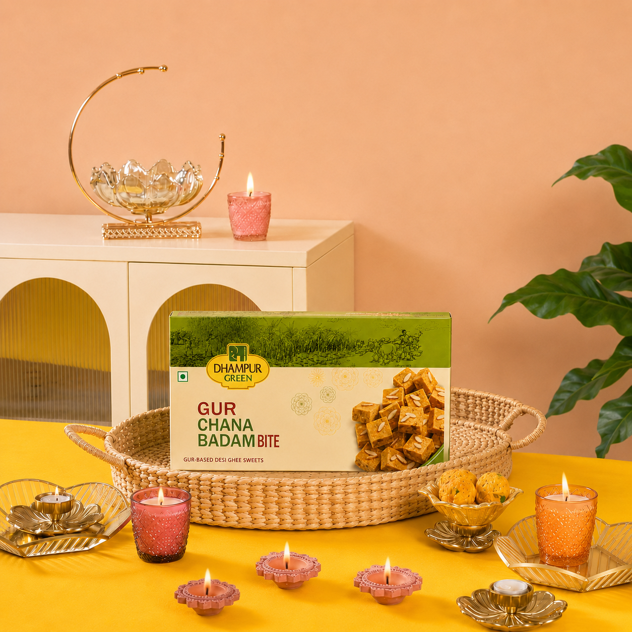 A box of Dhampur Green Gur Chana Badam Bite sweets is displayed in a woven tray on a yellow table, surrounded by pink and gold tea lights, with a matching styled background featuring a peach wall and white cabinet.