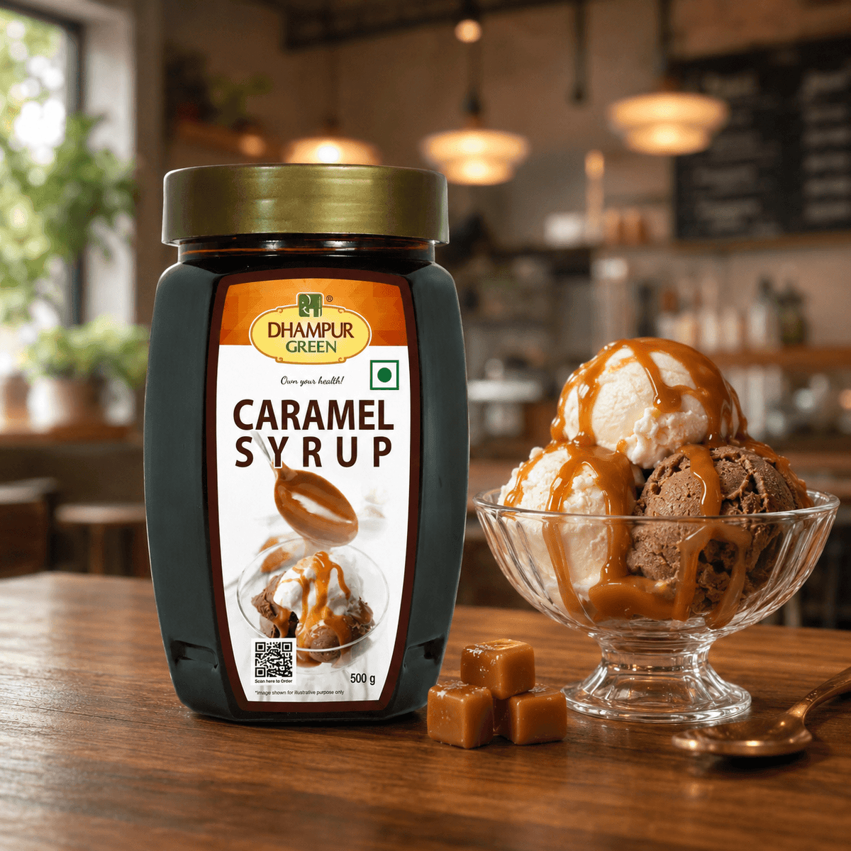 A jar of Dhampur Green Caramel Syrup stands on a wooden table beside a glass bowl of ice cream topped with caramel sauce and a few caramel cubes, with a softly blurred cafe interior in the background.