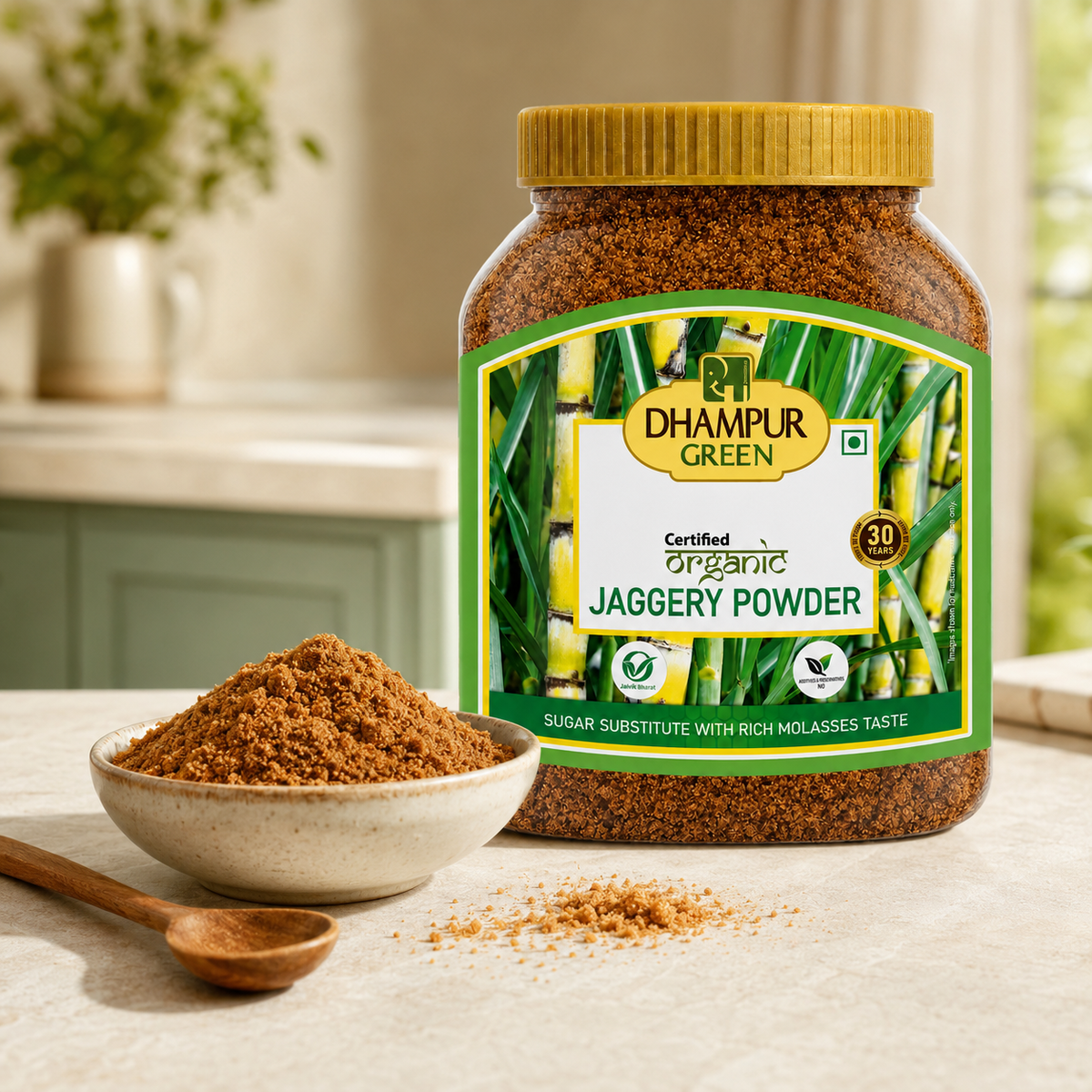 A jar of Dhampur Green Organic Jaggery Powder stands on a light countertop with a small bowl and wooden spoon of the brown powder in front. The background shows a softly blurred kitchen setting with green cabinets and a plant.