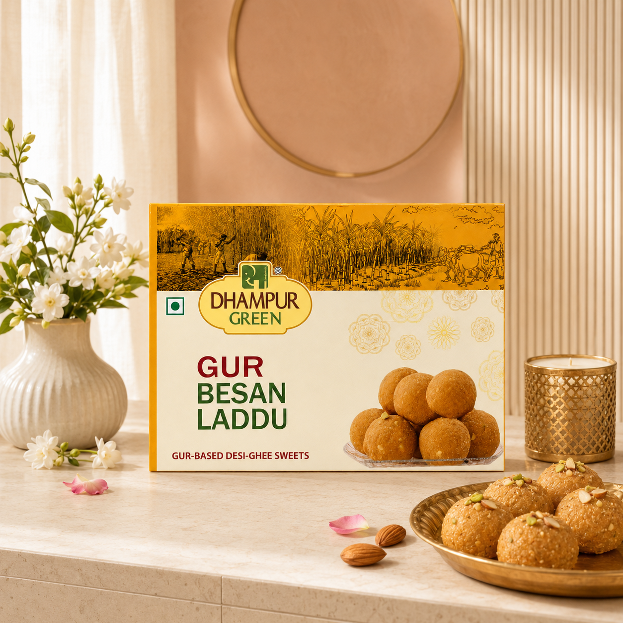 A box of Dhampur Green Gur Besan Laddu is displayed on a marble countertop with a plate of golden-brown laddus, almonds, and a decorative vase of white flowers in a warm, elegant setting.