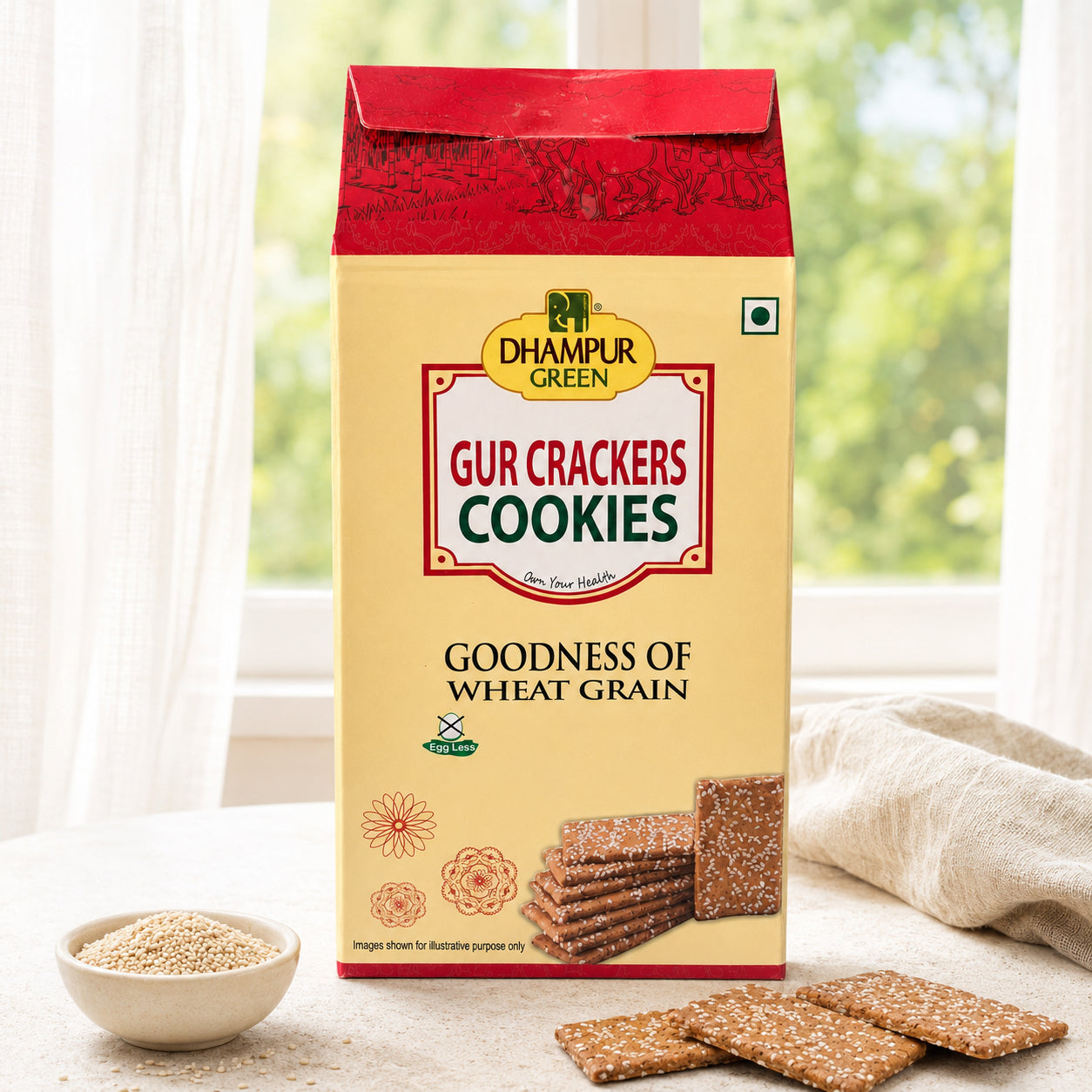 A package of Dhampur Green Gur Crackers Cookies in Goodness of Wheat Grain flavor, displayed on a table with a bowl of wheat flakes and several crackers in front, set against a soft-focus background of a window with curtains.