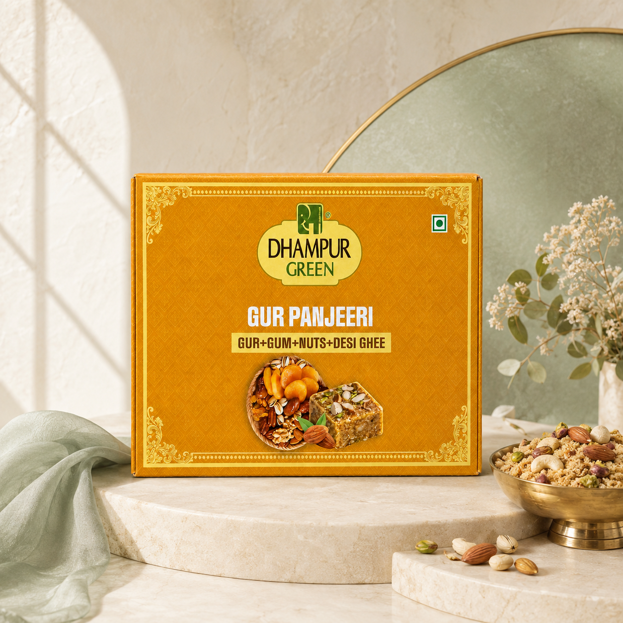 A box of Dhampur Green Gur Panjeeri (Gur + Gum + Nuts + Desi Ghee) displayed on a light beige marble pedestal. The packaging is a warm golden yellow with ornate gold borders and a photo of the prepared food on the front.