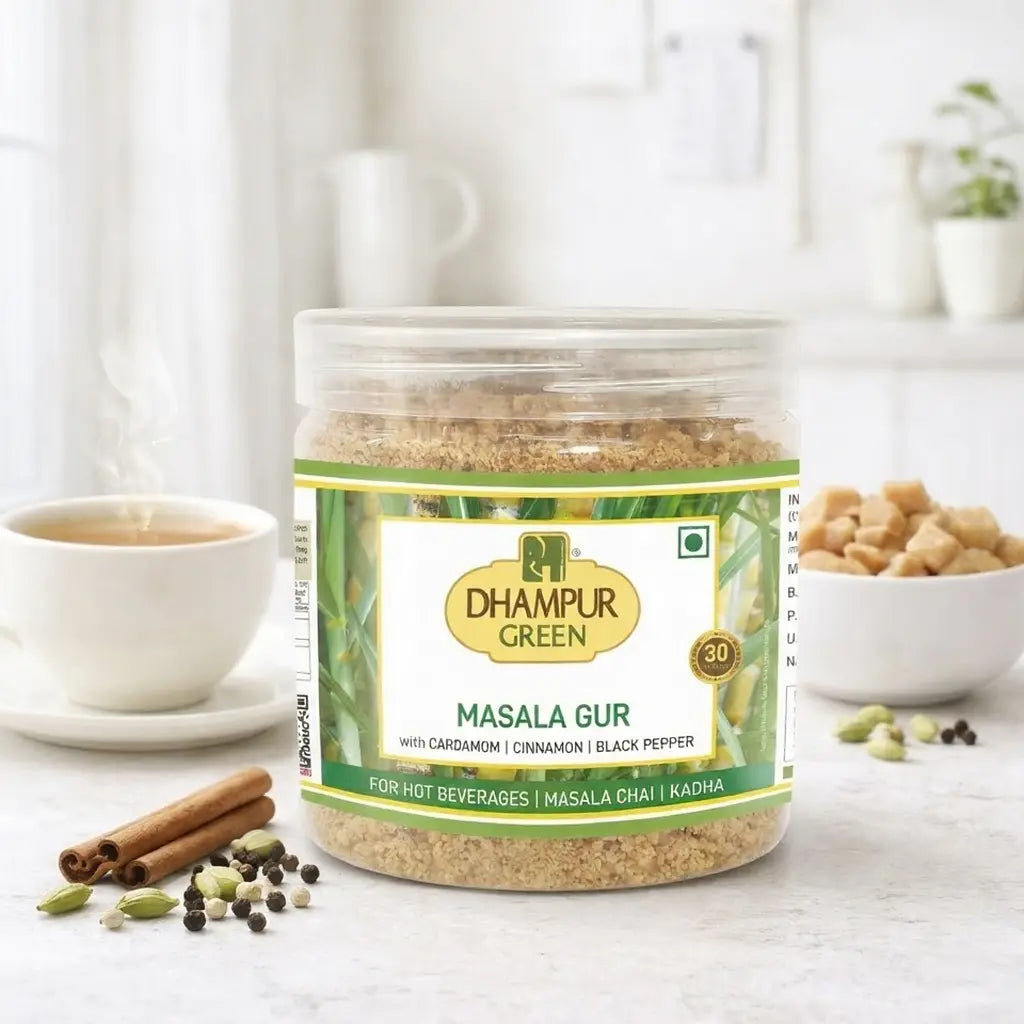 Jar of Dhampur Green Masala Gur on a white surface with a cup of tea and spices in the background.

