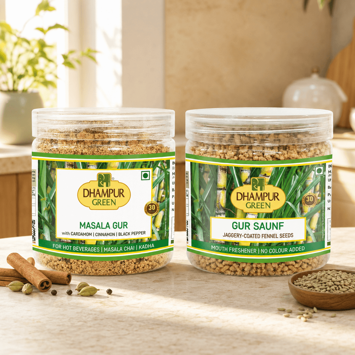 Two clear plastic spice jars labeled Dhampur Green Masala Gur and Gur Saunf, displayed on a light countertop with cinnamon sticks, green cardamom pods, and a small bowl of seeds. The background shows a softly blurred kitchen setting.