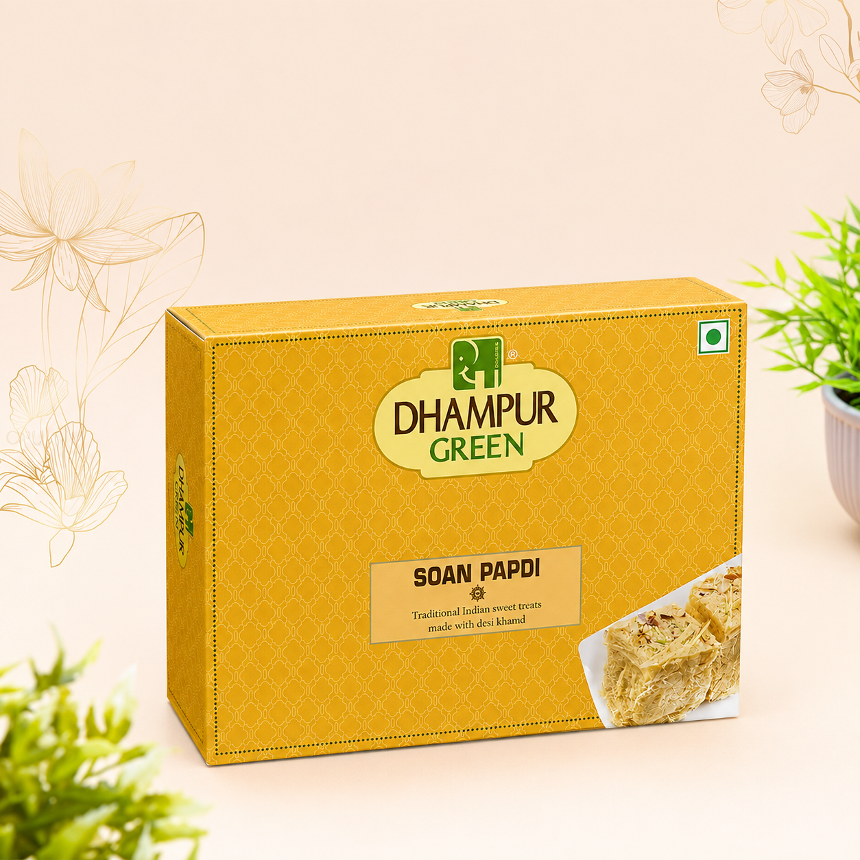 A box of Dhampur Green Soan Papdi, featuring a golden-yellow patterned design with the brand logo in the center and a small image of the sweet pastry on the bottom right. The box is styled on a light beige background with decorative floral line drawings and small potted plants.